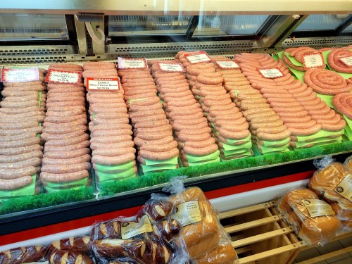 A display of various sausages in a butcher shop, with some packaged bread rolls in the foreground.