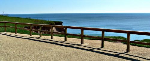 A wooden railing along a sandy path overlooking a calm blue sea under a clear sky.
