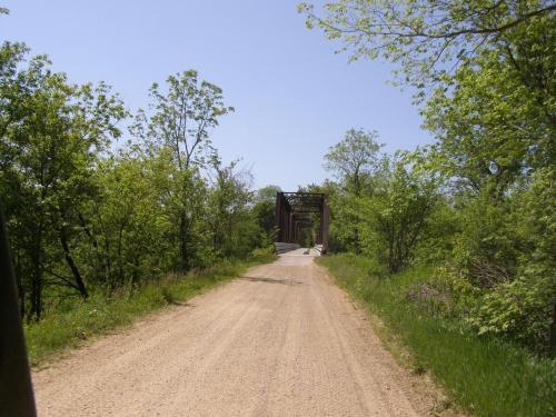 A dirt road leads to a metal bridge surrounded by lush green trees under a clear blue sky.