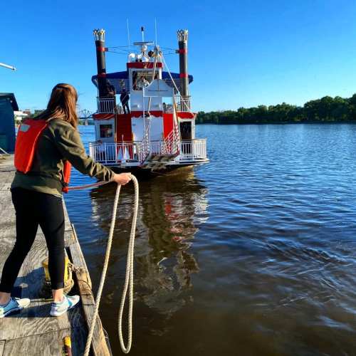 A person in a life jacket pulls a rope towards a colorful riverboat on a calm waterway under a clear blue sky.