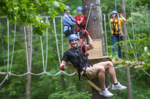 A person zip-lining through a forest, smiling, with others in the background on a ropes course.