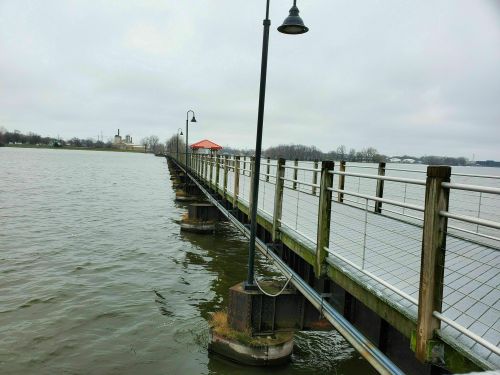 A long pier extends over calm water, with lampposts and a red-roofed shelter in the distance under a cloudy sky.