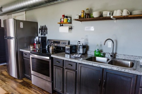 Modern kitchen with stainless steel appliances, granite countertops, and open shelving displaying dishes and condiments.