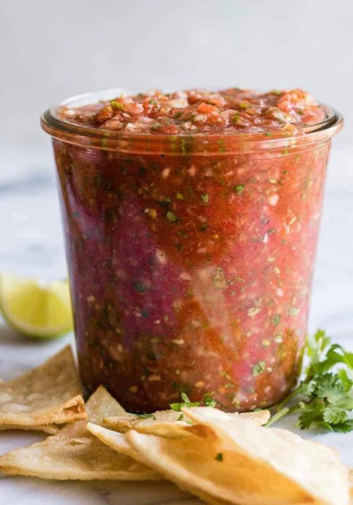 A jar of fresh salsa surrounded by tortilla chips and a lime wedge on a marble surface.