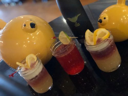 Three colorful cocktails with lemon slices, alongside two yellow decorative birds and a bottle in the background.