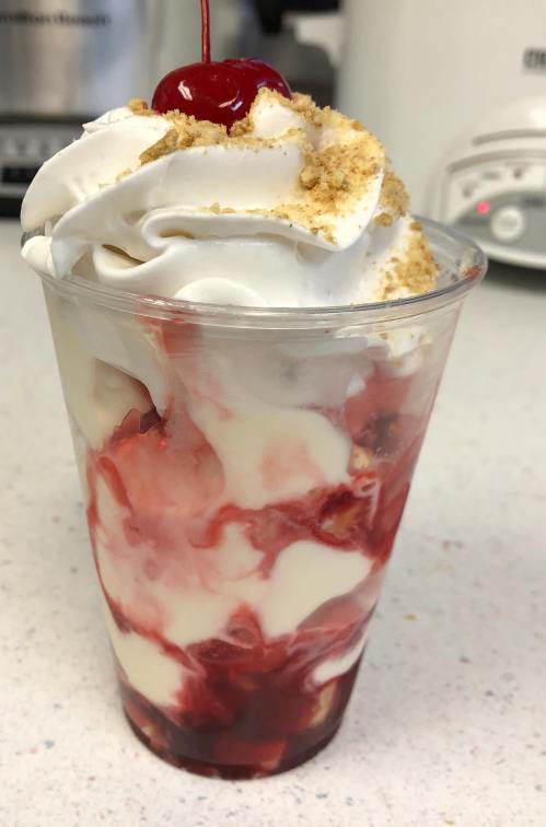 A delicious dessert in a clear cup, featuring layers of whipped cream, cherry topping, and crumbled graham crackers.