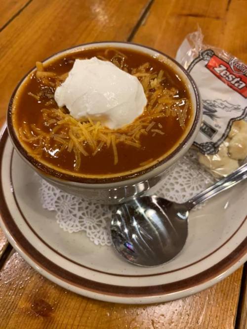 A bowl of chili topped with shredded cheese and sour cream, served with a spoon and a small packet of crackers.