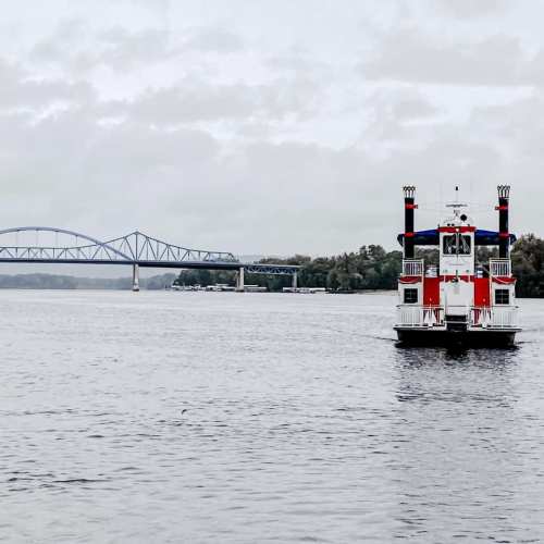 A red and white riverboat sails on a calm river, with a bridge visible in the background under a cloudy sky.