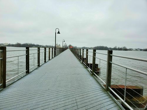 A long wooden pier extends over a calm body of water, with lampposts lining the sides under a cloudy sky.