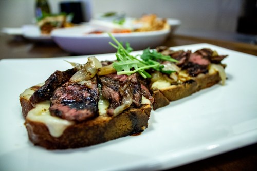 A close-up of a gourmet sandwich with grilled steak, melted cheese, and arugula on toasted bread, served on a white plate.