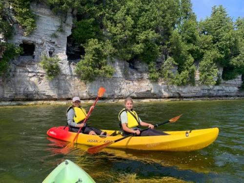 Two people kayaking in bright yellow and red kayaks near a rocky shoreline with lush greenery above.