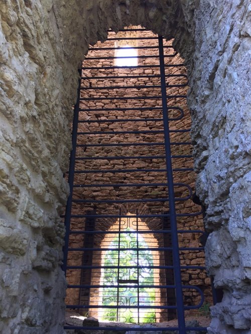 View through a stone archway with a wrought iron gate, revealing a bright window and greenery beyond.