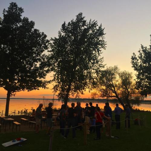 A group of people gathered by a lake at sunset, with trees silhouetted against a colorful sky.