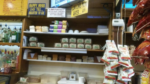 A bar shelf displaying various snacks, drinks, and packaged food items, with a TV and signs in the background.