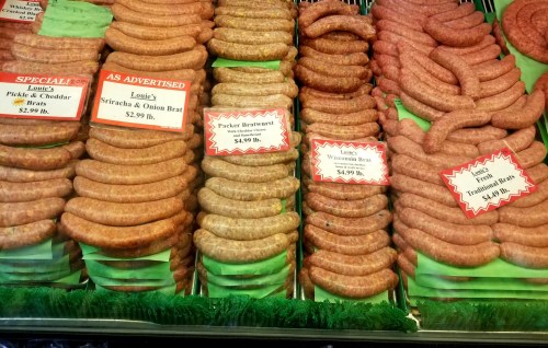 Display of various types of sausages, including pickle & cheddar, sriracha & onion, and traditional bratwurst, with prices.