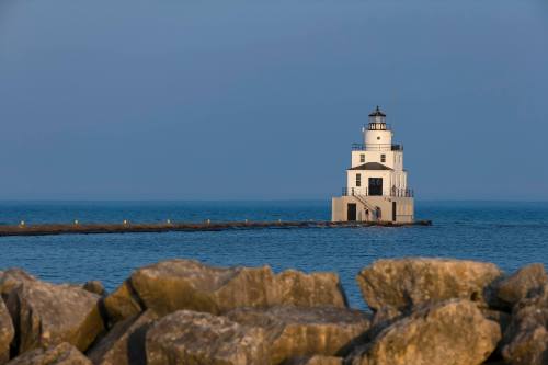 A white lighthouse stands on a pier, surrounded by calm blue waters and rocky shoreline under a clear sky.