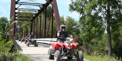 ATVs crossing a metal bridge surrounded by greenery on a sunny day.