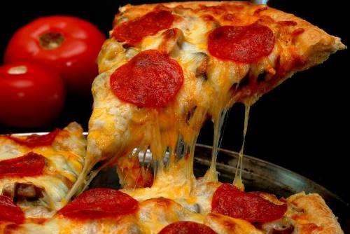 A slice of pepperoni pizza with melted cheese, being lifted from a pan, with fresh tomatoes in the background.