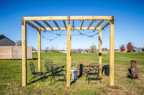 A wooden pergola with string lights, surrounded by chairs and a small table on a grassy area.