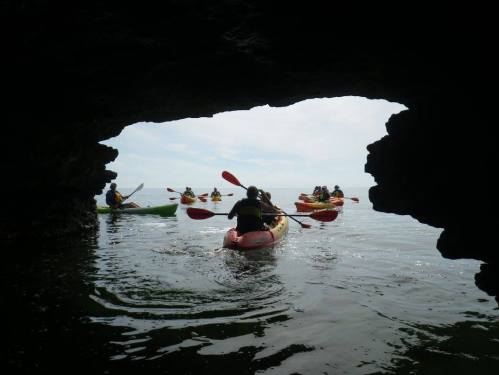 Kayakers paddle through a rocky archway, exploring calm waters under a cloudy sky.
