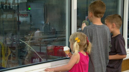 Three children watch a factory process through a window, holding ice cream cones, with machinery visible in the background.