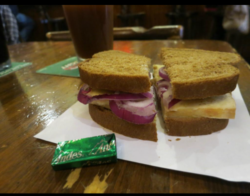 A sandwich with layers of bread, chicken, and red onion, next to a green Andes mint on a wooden table.