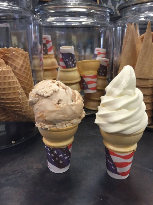 Two ice cream cones with patriotic wrappers: one with a scoop of brown ice cream and the other with soft-serve vanilla.