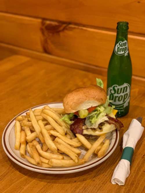 A plate of golden fries next to a burger topped with lettuce, pickles, and bacon, with a bottle of Sun Drop soda.