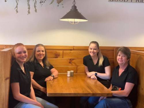Four women sit together at a wooden table in a cozy restaurant, smiling and wearing matching black shirts.