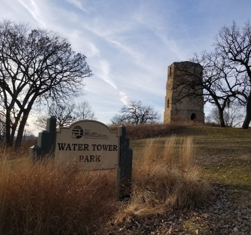 A sign for Water Tower Park with a historic stone tower and bare trees in the background under a blue sky.