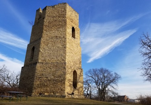 A tall, weathered stone tower stands against a blue sky with wispy clouds, surrounded by trees and grassy land.
