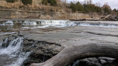 A serene waterfall cascades over rocky ledges, surrounded by trees and a calm river.