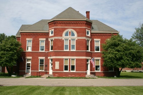 Historic red brick building with a symmetrical design, large windows, and American flags, surrounded by green trees.
