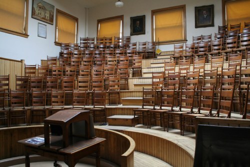 An empty lecture hall with wooden chairs arranged in a semicircle and a small desk in the foreground.