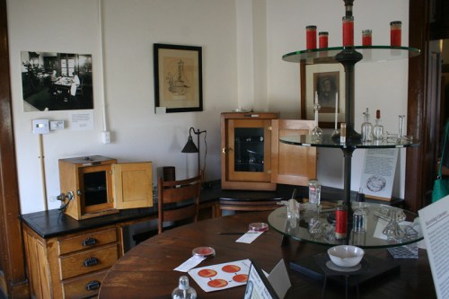 A vintage laboratory setup with glassware, a wooden cabinet, and historical photographs on the walls.