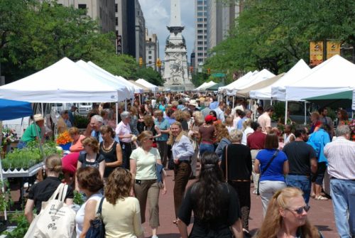 A bustling outdoor market with many people browsing tents filled with plants and flowers, set against a city backdrop.