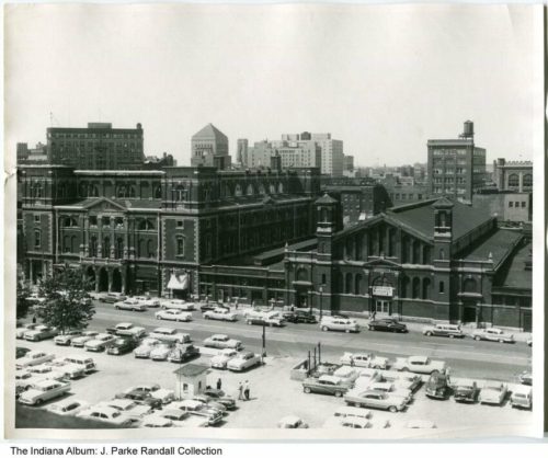 Aerial view of a city block featuring historic buildings and a parking lot filled with vintage cars.