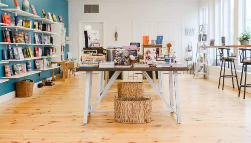 A cozy bookstore interior featuring a wooden table with books and decor, surrounded by shelves and natural light.