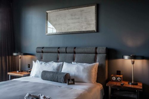 A modern hotel room featuring a gray upholstered bed, bedside tables with lamps, and dark blue walls.