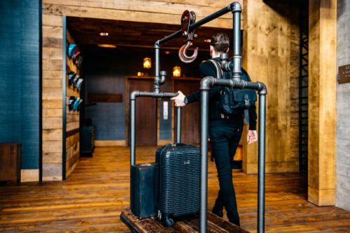 A person walks away with a suitcase on a luggage cart made of pipes in a rustic hotel lobby.