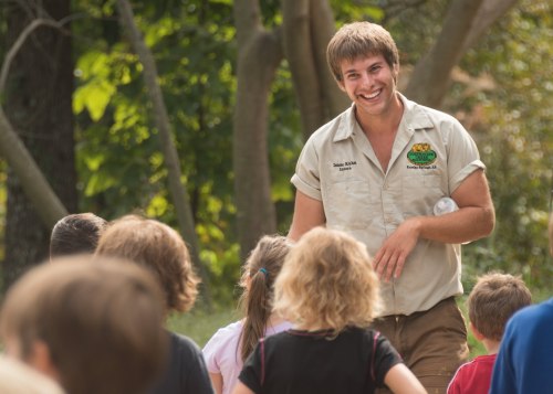 A smiling guide interacts with a group of children in a natural outdoor setting.