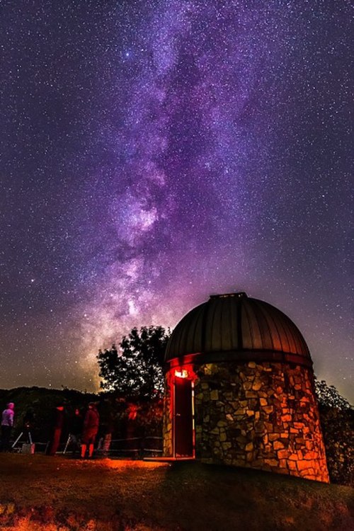 A stone observatory under a starry sky, with the Milky Way visible and people stargazing nearby.