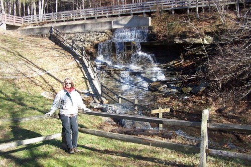 A person stands by a small waterfall, surrounded by greenery and wooden fencing in a serene outdoor setting.