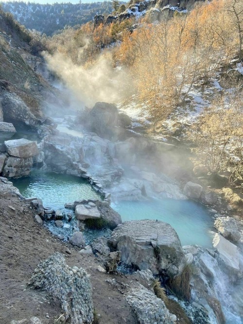 A serene landscape featuring steaming hot springs surrounded by rocky terrain and autumn trees.