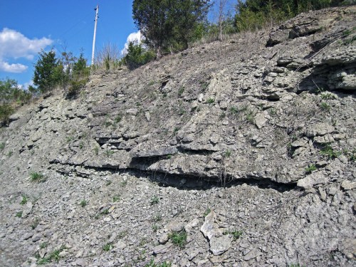 A rocky hillside with layered sedimentary stones and sparse vegetation under a blue sky with scattered clouds.