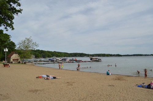 A sandy beach with people swimming and relaxing, boats docked in the background, under a cloudy sky.