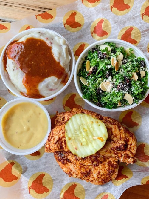 A plate featuring grilled chicken with a pickle, mashed potatoes with gravy, a kale salad, and a side of cheese sauce.