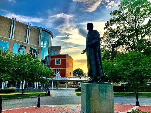 A statue of a figure stands in a park, surrounded by trees and modern buildings under a colorful sunset sky.