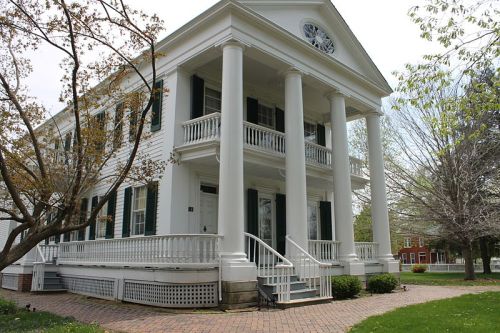 A white, columned building with green shutters, surrounded by trees and a brick pathway.