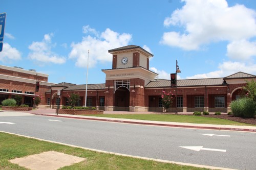 Exterior view of a brick high school building with a clock tower, surrounded by greenery and a blue sky.